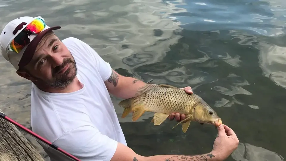 A man with tattoos holds a small carp by a lake, showcasing a casual, triumphant expression. He wears a white shirt and cap, holding a fishing rod.