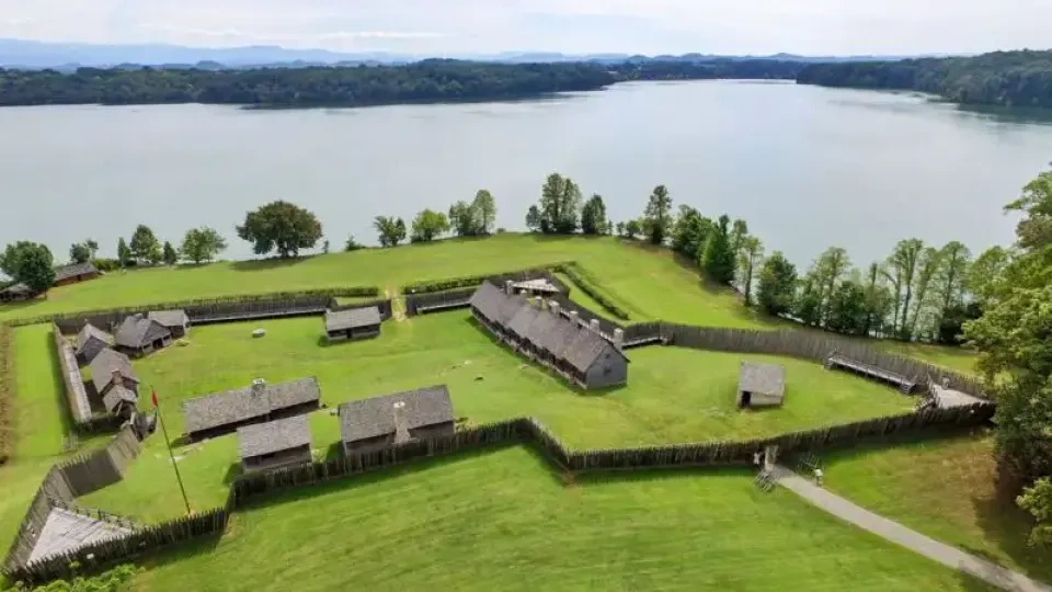 Aerial view of a historic fort by a lake, featuring a large wooden structure and smaller huts enclosed by a palisade, surrounded by lush greenery.