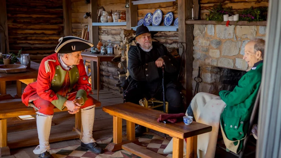 Three men in historical attire converse inside a rustic cabin, surrounded by wooden furniture and blue plates on shelves, creating a nostalgic atmosphere.