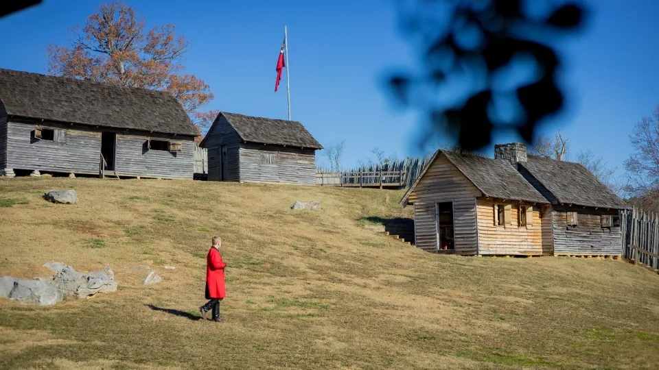A person in a bright red coat walks on a grassy hill in front of rustic wooden cabins under a clear blue sky, conveying a sense of serenity and history.