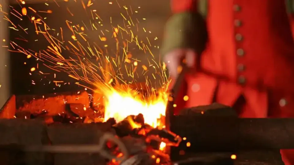 A vibrant forge scene with glowing red coals and bright orange sparks flying. A blurred figure in red and green tends the fire, creating a warm, dynamic atmosphere.