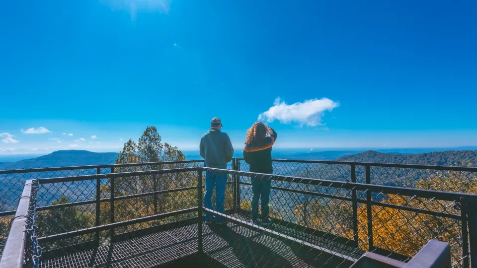 Two people stand on a metal viewing platform, gazing at a vast landscape of rolling hills under a clear blue sky with the sun shining brightly.