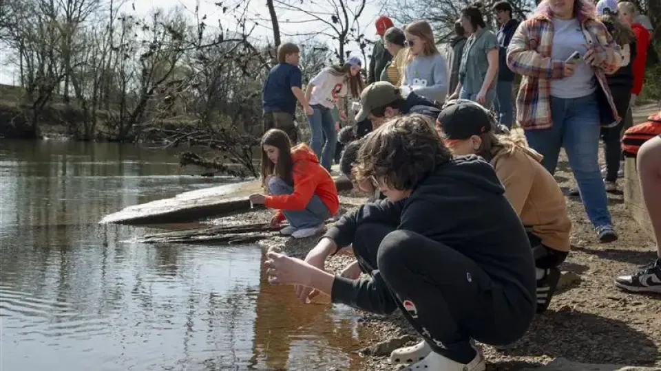 Group of people gathered by a riverbank, some squatting and touching the water.