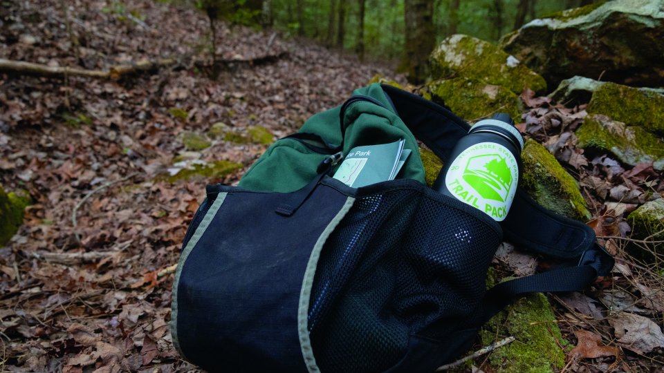 A green backpack with a "National Park" tag sits on a forest trail, containing a water bottle and journal. 