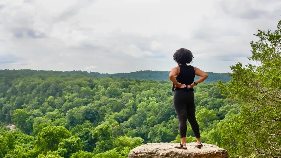 A person stands confidently on a rock ledge, overlooking a lush, green forest under a cloudy sky. The scene conveys a sense of adventure and tranquility.
