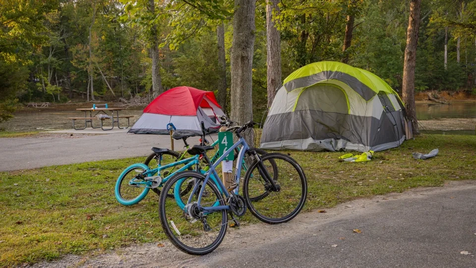 Two tents, one red and gray, the other green and gray, are set up by a lakeside. Three bikes are parked nearby, and autumn trees surround the campsite.