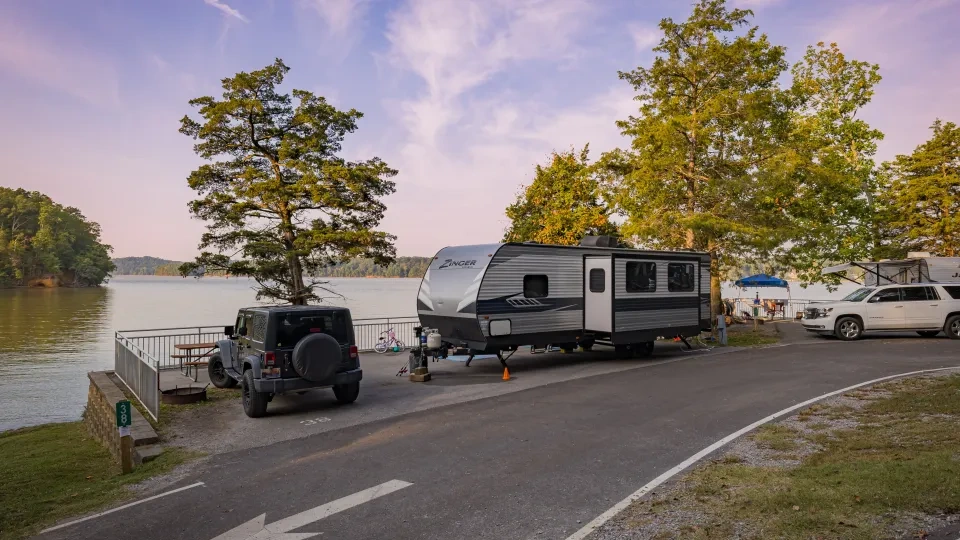 A serene lakeside campsite at sunset, featuring a parked RV next to a tree and a Jeep. The sky is pinkish-purple, and there's a calm, tranquil vibe.