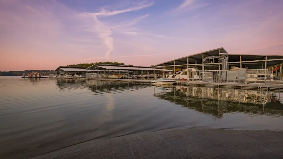 A serene marina at sunset with docks and covered boats reflected on calm water. The sky is tinged with pink and purple hues, creating a tranquil atmosphere.