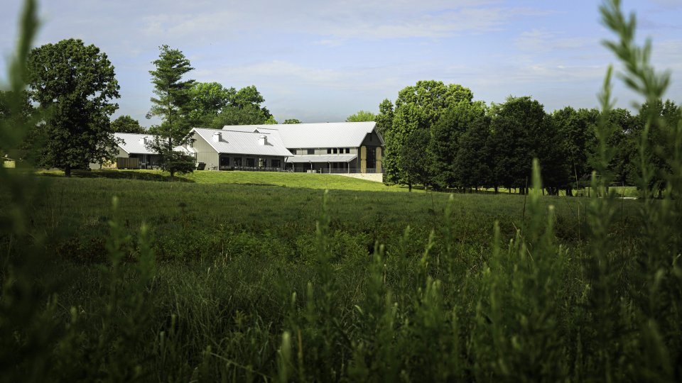 A modern lodge-style building with a metal roof surrounded by open grassy fields and trees under a blue sky.