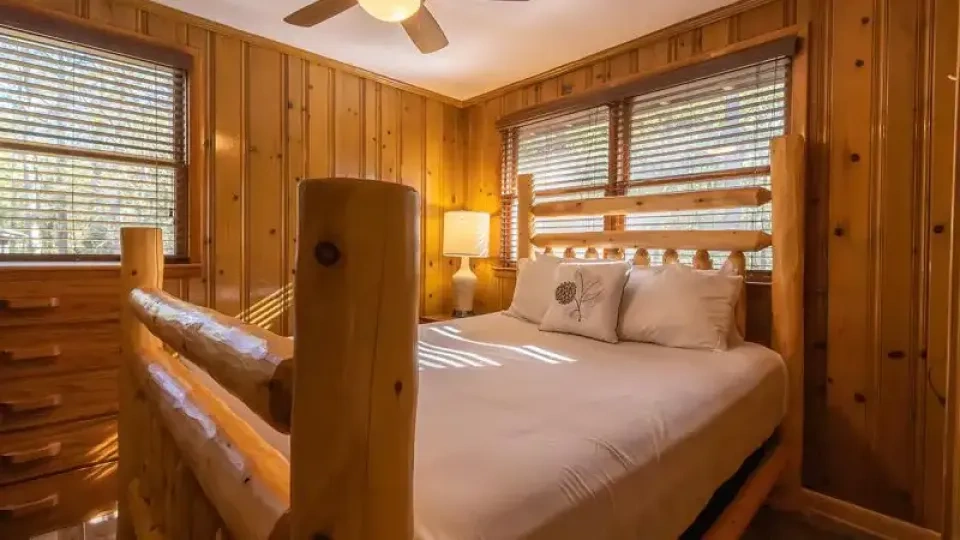 Bedroom with wood-paneled walls, a rustic log-frame bed, and sunlight streaming through two windows.