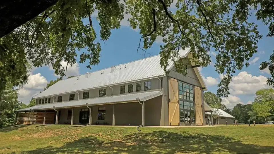 A modern barn with large glass windows and a sloped metal roof stands under a tall tree against a blue sky with clouds, evoking a serene rural atmosphere.