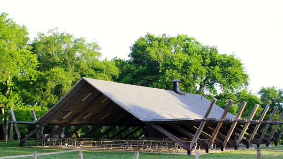 A wooden pavilion with a large peaked roof and many picnic tables set in a lush green park. Sunlight casts shadows, creating a serene and inviting outdoor setting.
