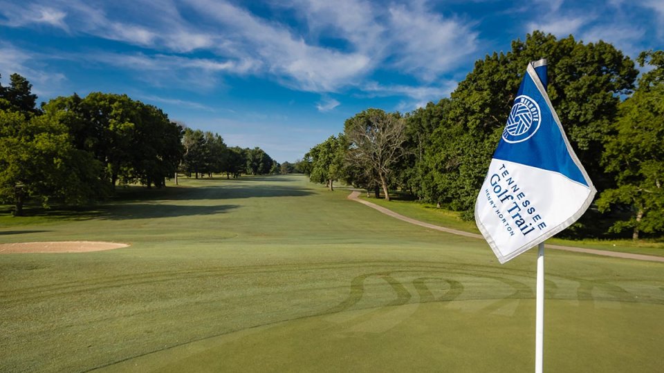 A golf course with a blue sky and scattered clouds overhead. In the foreground, a flag labeled "Tennessee Golf Trail" flutters near a sand trap.