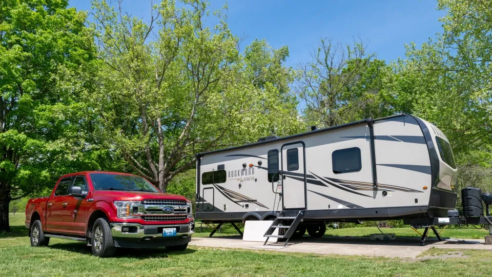Red truck and RV trailer parked on grass under trees, sunny day.
