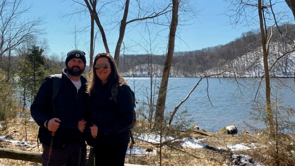 A couple stands smiling with hiking poles beside a snowy lake on a sunny winter day. Bare trees frame the scene, conveying a peaceful, chilly ambiance.