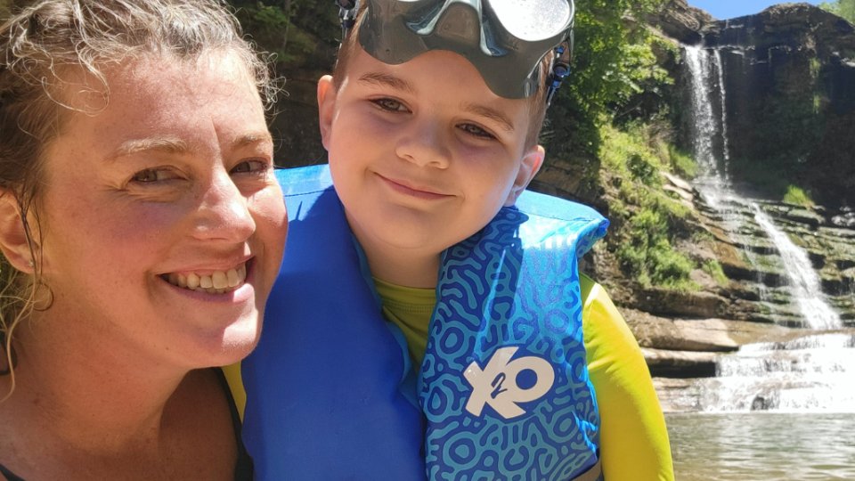 A smiling woman and child wearing a blue life vest pose for a selfie in front of a cascading waterfall in lush greenery under a bright, sunny sky.