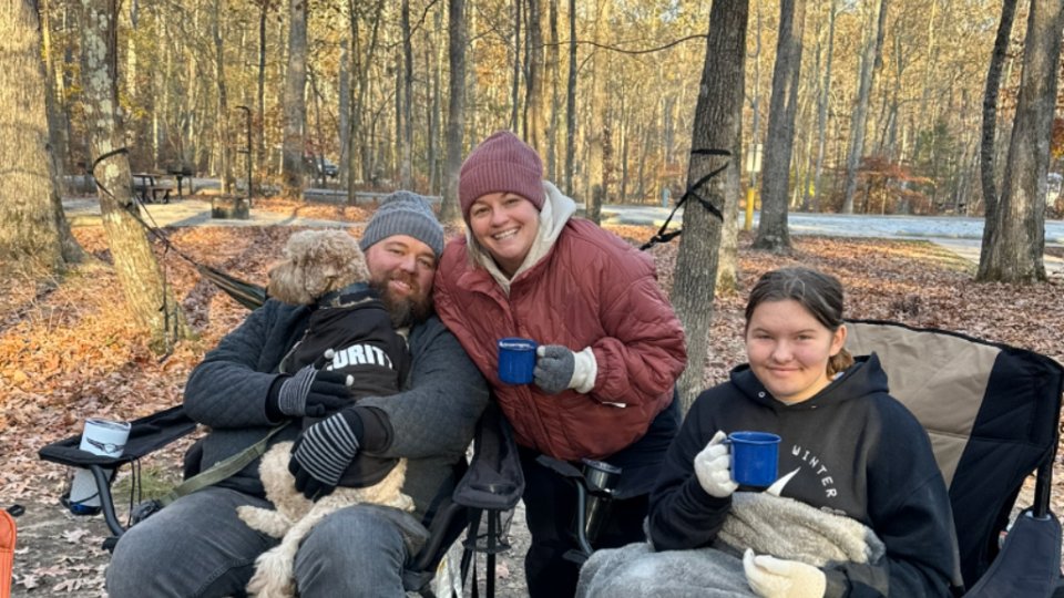 A family enjoys a cozy outdoor setting, sipping from blue mugs. They sit in chairs among autumn trees with warm clothes and smiles.