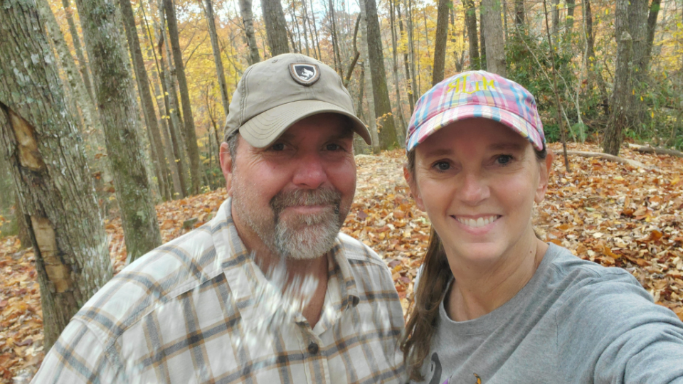 A smiling man and woman, wearing caps, take a selfie in a forest during autumn. Fallen leaves cover the ground, and trees with orange leaves create a warm backdrop.