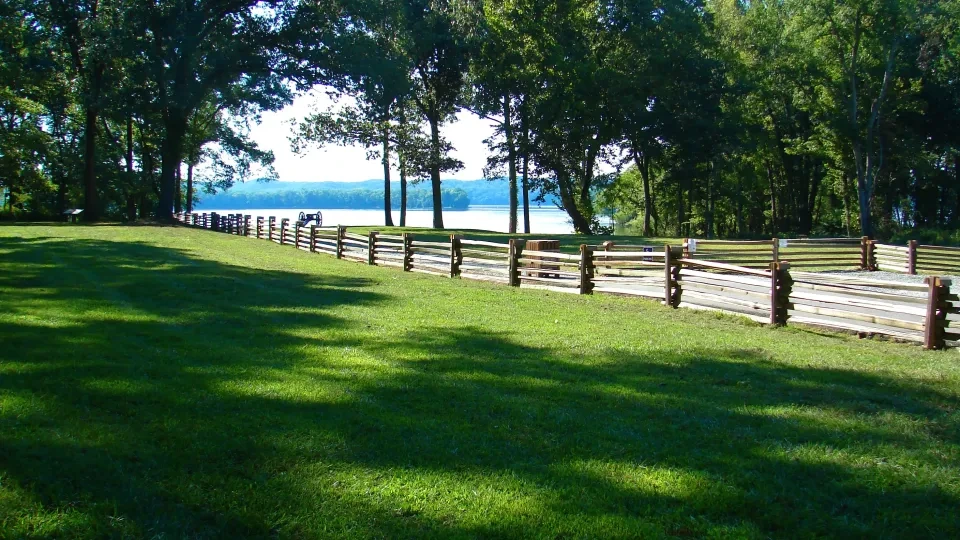 Sunny park with wooden fence, green grass, and trees by a lake.