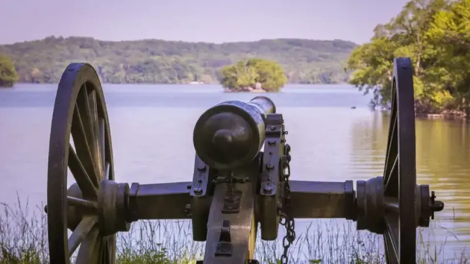 Cannon aimed over a calm lake, with trees in the distance.