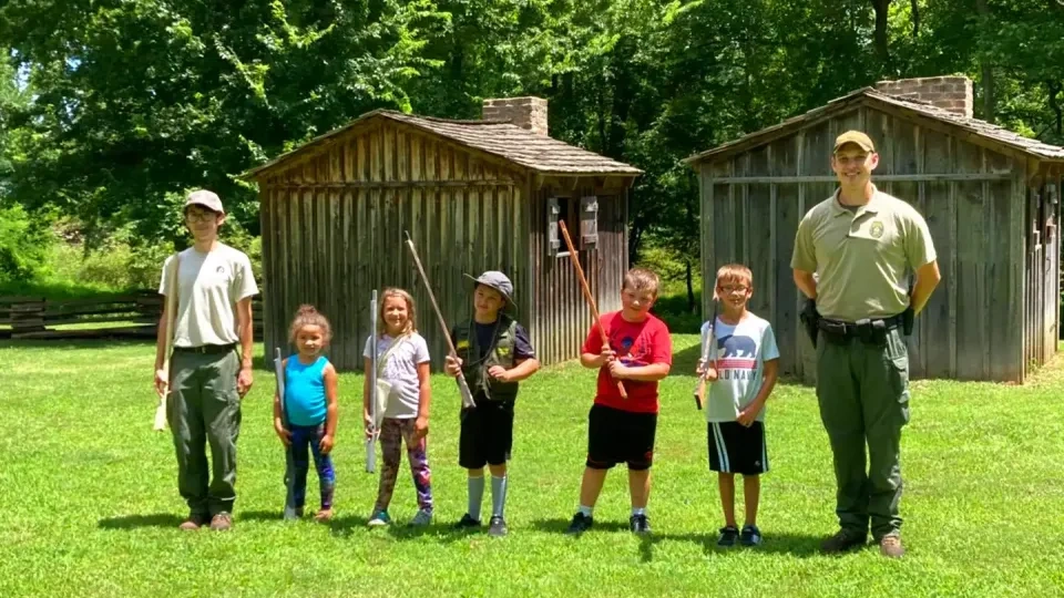 Group of kids with sticks and two adults standing on grass near wooden cabins.