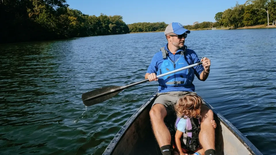 A man and a child in life jackets paddle a canoe on a tranquil lake. Surrounded by lush green trees, the scene conveys a peaceful, sunny day.
