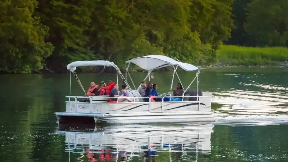 A group of people are enjoying a leisurely ride on a white pontoon boat. The boat glides across a calm lake, surrounded by lush green trees under overcast skies.