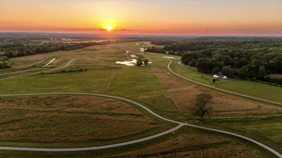 Aerial view of a serene landscape at sunset, with the sun setting over green fields, winding roads, a small pond