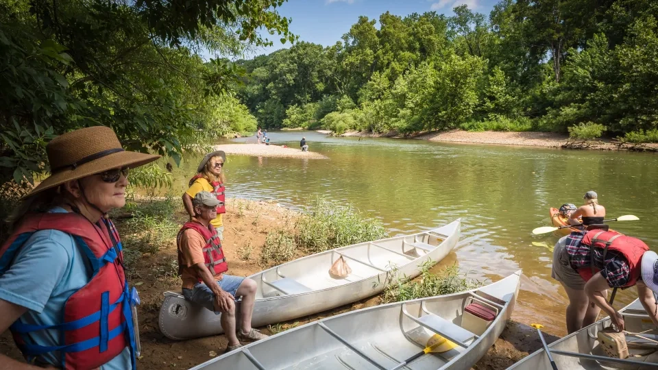 Canoeists in life vests by a riverbank, surrounded by greenery, with canoes on the shore.