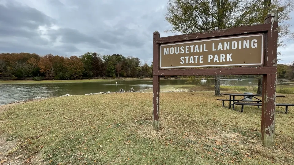 A wooden park sign reading “Mousetail Landing State Park” stands on a grassy area near the water’s edge.