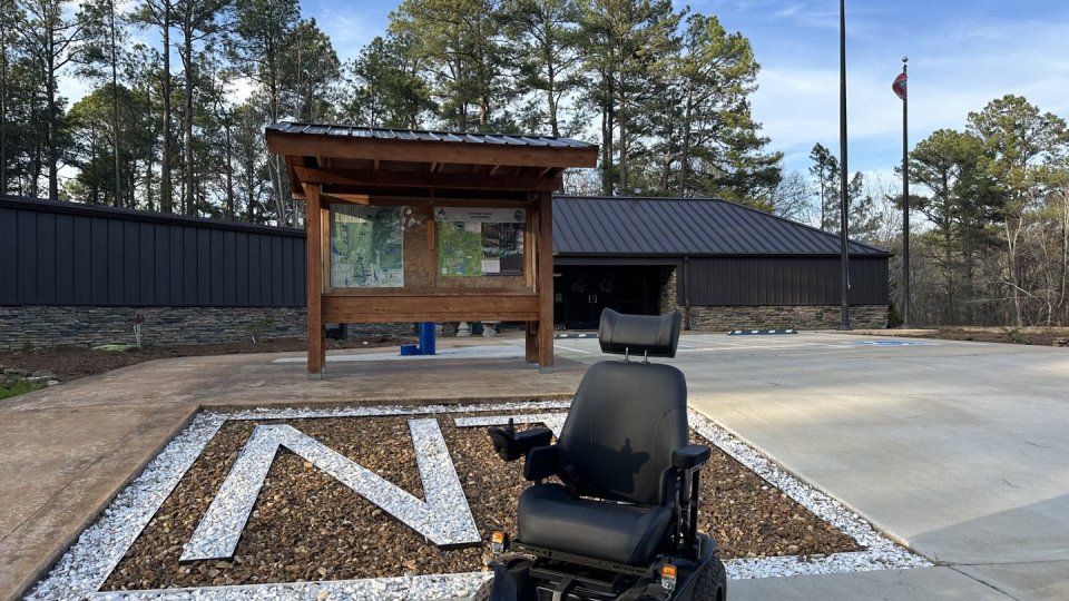 A black electric wheelchair is parked near a wooden information board outside a building, with a paved area featuring a large "N". 