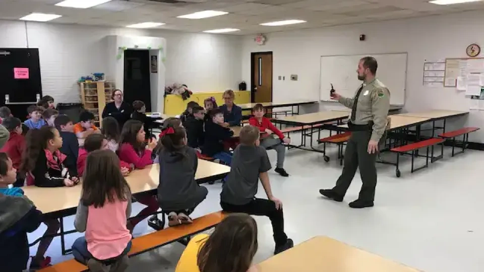 Man in uniform speaking to children seated at cafeteria tables.