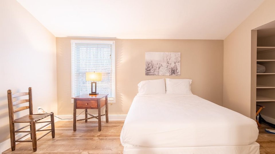 Bright bedroom with one bed, a wooden chairs, nightstand with a lamp on top, and a window