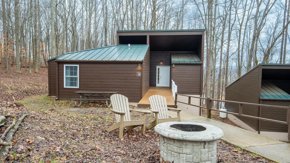 Brown cabin with green roof with stone fire bit and chairs in front, has wooden bridge leading to door