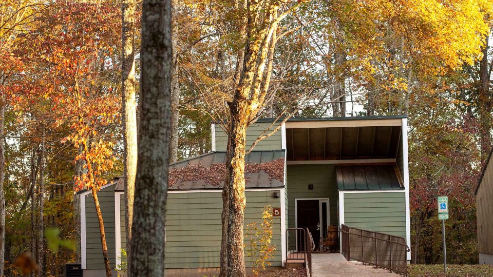 Green cabin surrounded by trees with a bridge leading to the front door located next to handicap parking