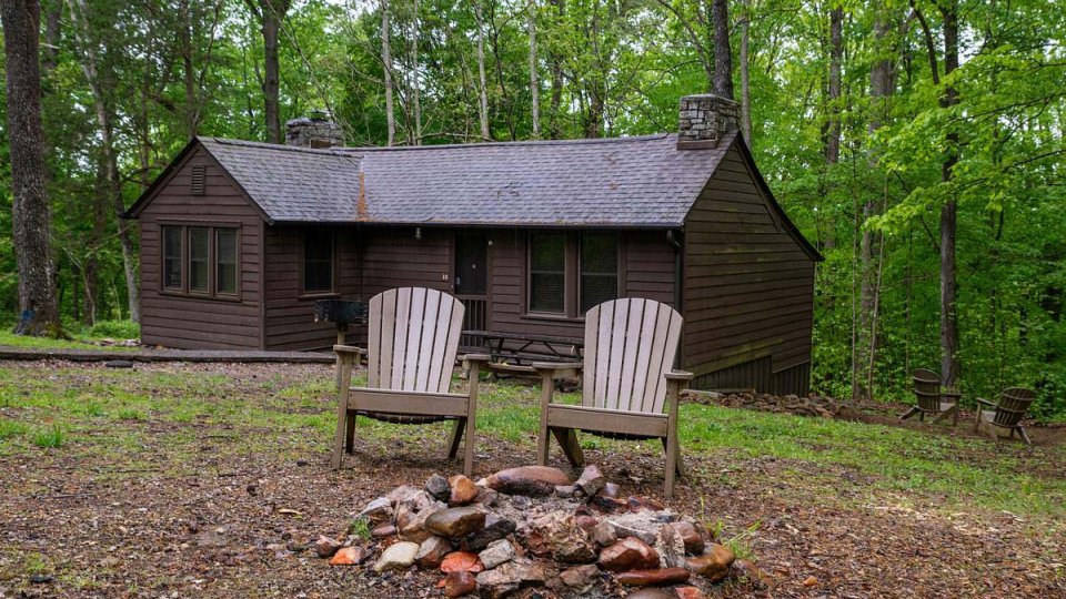 Two Adirondack chairs by a fire pit, in front of a rustic cabin in the woods.