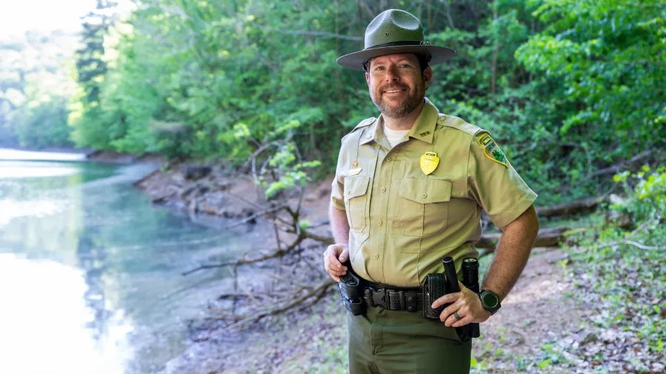 Park ranger smiling by a forested riverbank wearing a uniform and hat.