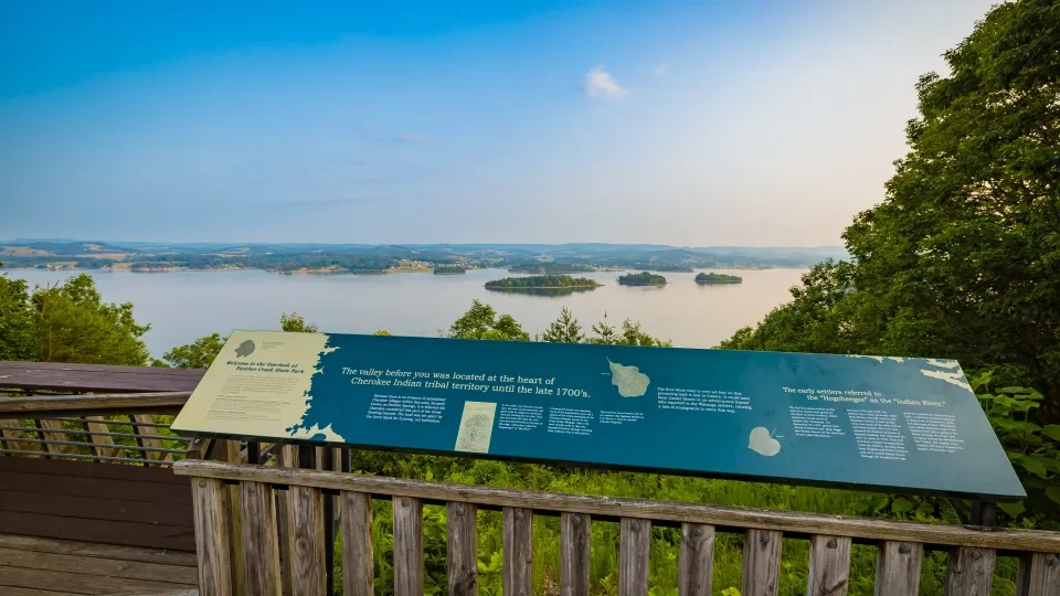 Overlook with sign and view of serene lake and distant hills under a clear blue sky.