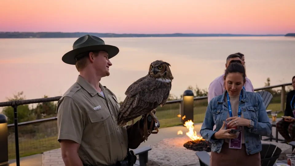 a park ranger, holding a large screech owl, with a beautiful sunset is in the background over the lake. 