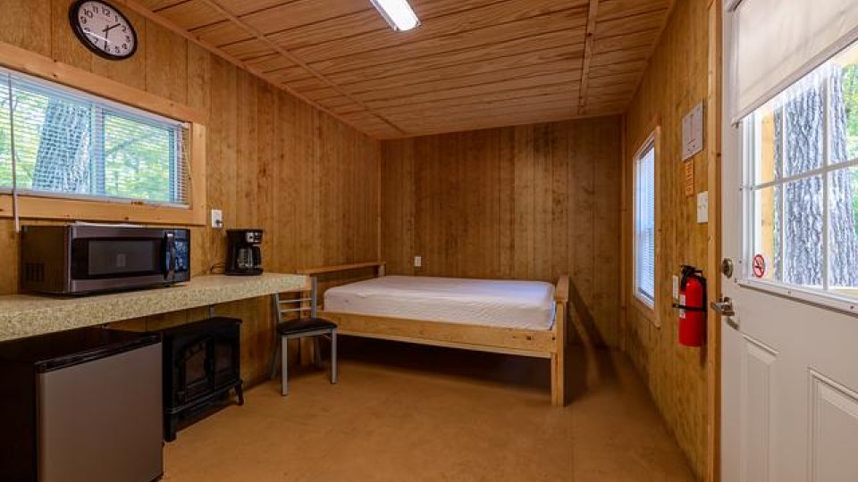 Wooden room with over head lighting, bed, windows, door, fire extinguisher, mini fridge, microwave, chair, and coffee maker