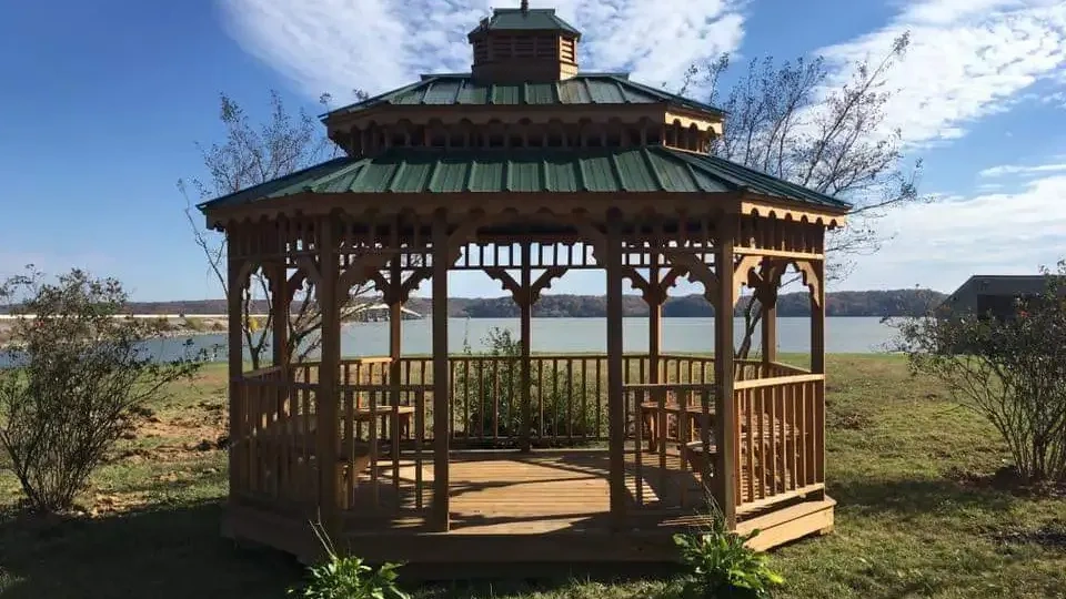 Wooden gazebo with a green roof by a lake under a cloudy sky.