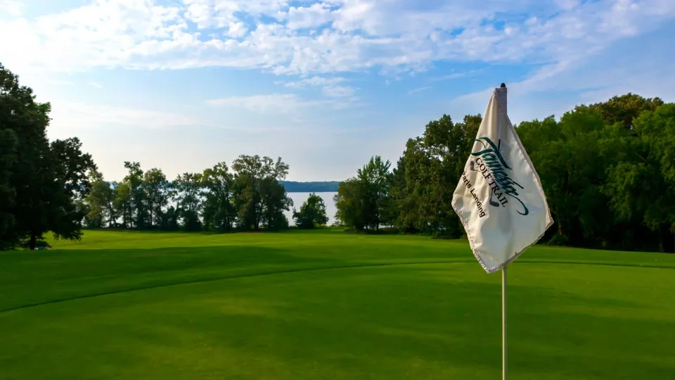 Golf course with flag, trees, and a lake under a partly cloudy sky.