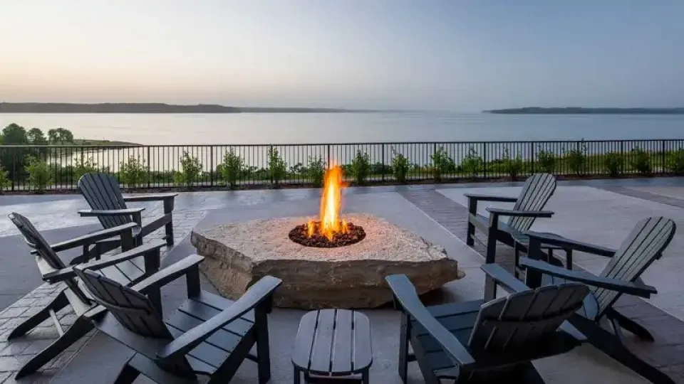 Fire pit with chairs on a patio overlooking a calm lake at sunset.