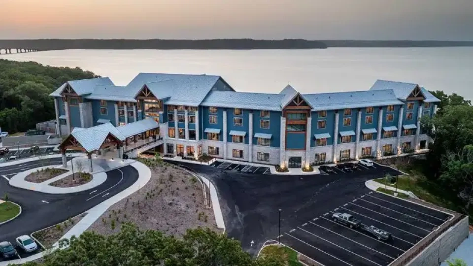 Large lakeside hotel with blue facade, surrounded by greenery at sunset.