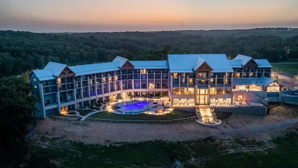 Hotel exterior at dusk with lit windows, pool, and surrounding forest.