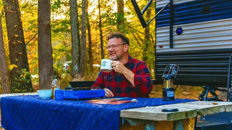 Man in plaid shirt enjoys coffee at a campsite, surrounded by autumn trees and a camper.