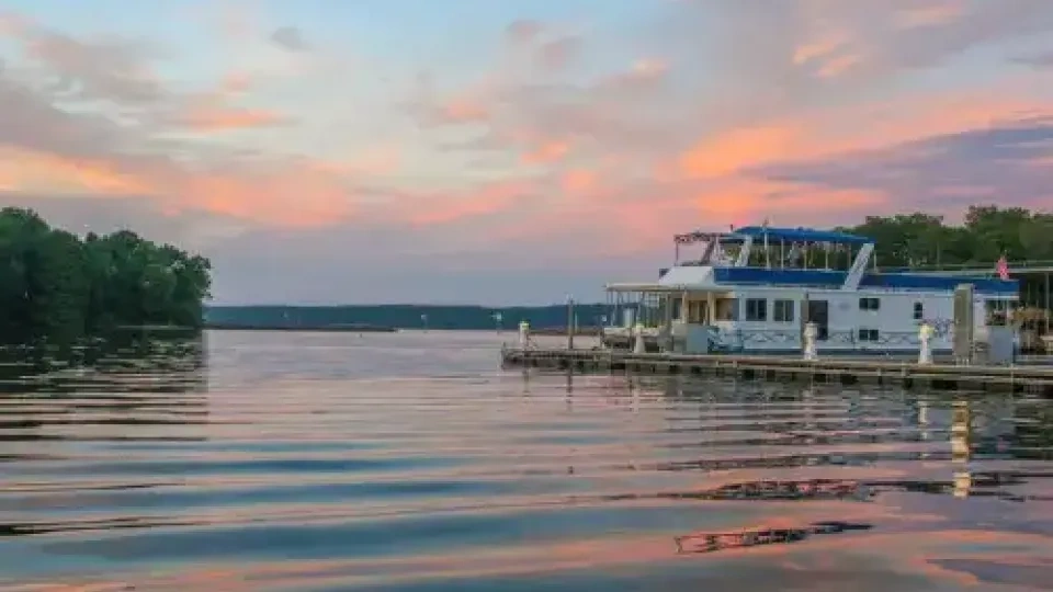 Houseboat docked on a calm lake at sunset with pink clouds.