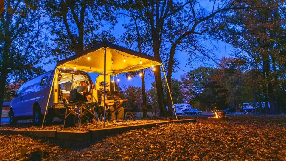 Van campsite at dusk, warm lights, people playing guitar, surrounded by autumn trees.