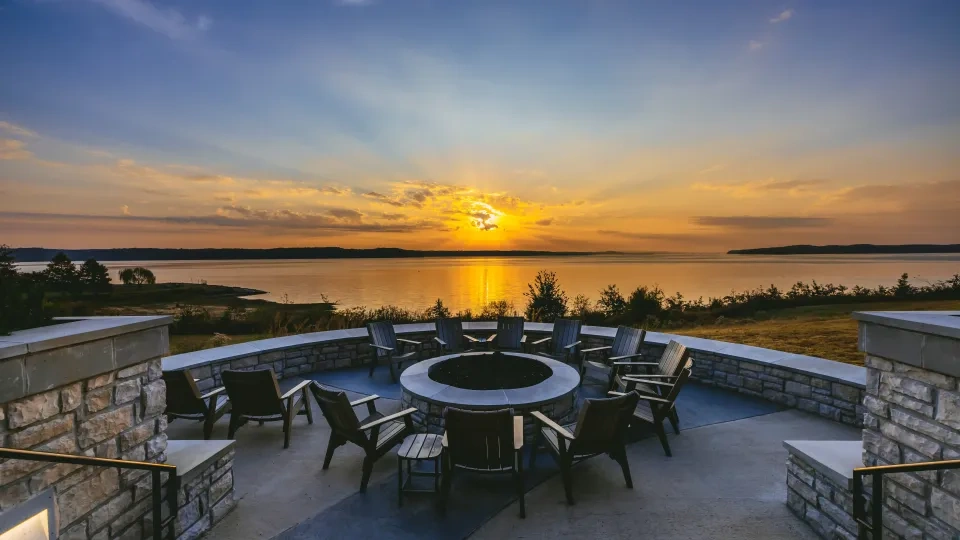 Sunset over lake with stone fire pit and chairs in foreground.