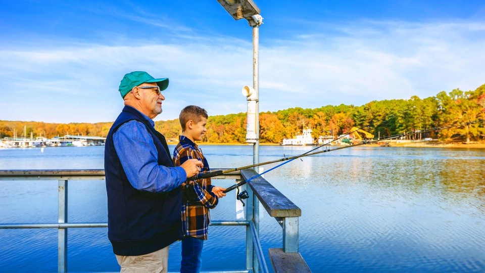Man and boy fishing on a lakeside pier, surrounded by autumn trees.